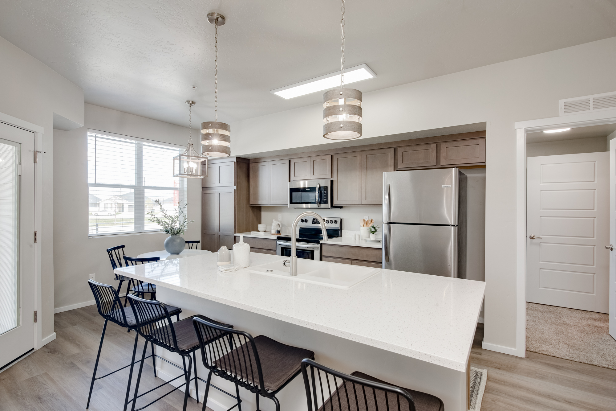 Modern open-concept kitchen featuring a spacious white quartz island with a built-in sink, black barstools, stainless steel appliances, and warm wood cabinetry. Pendant lighting adds elegance, while a cozy dining area with natural light offers a welcoming space for gatherings. A stylish and functional home covered by the 2-10 home buyers warranty.