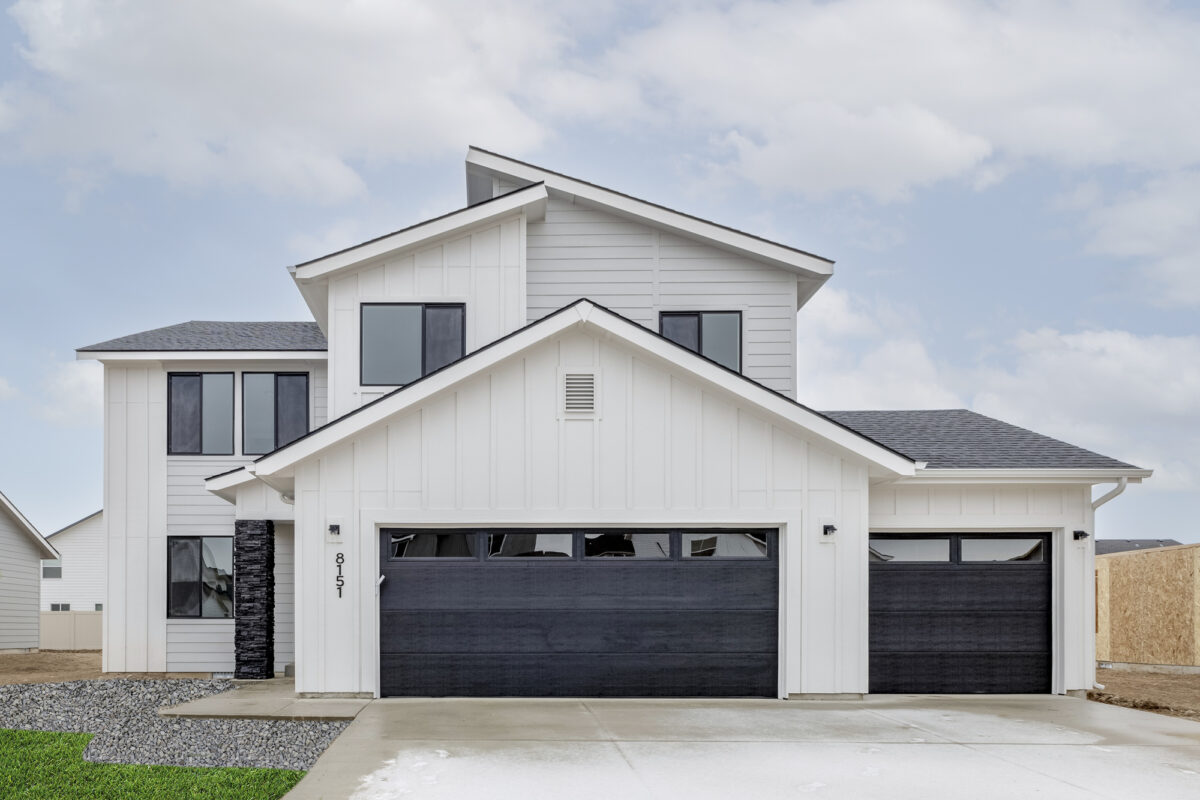 Modern white two-story home with black garage doors, large windows, and a covered entryway, set against a cloudy sky. This newly built home showcases contemporary farmhouse design, highlighting quality craftsmanship and protection with a 2-10 home buyers warranty.