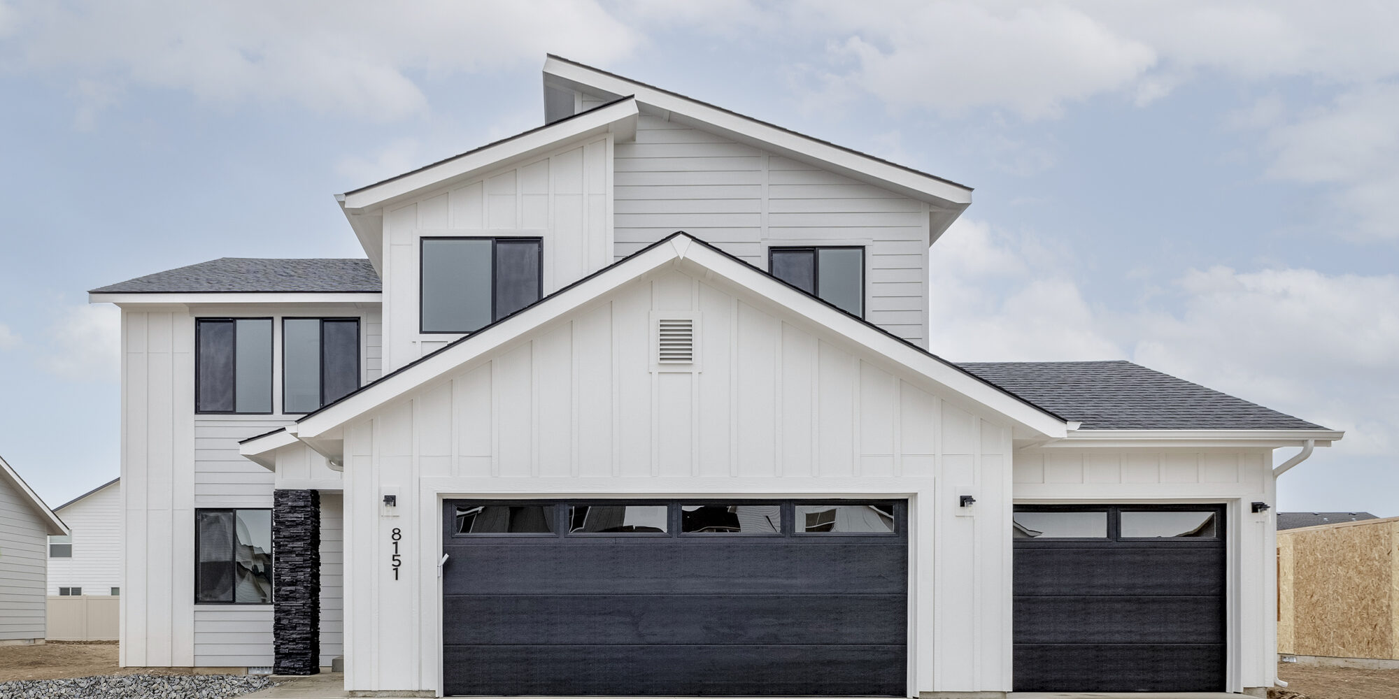Modern white two-story home with black garage doors, large windows, and a covered entryway, set against a cloudy sky. This newly built home showcases contemporary farmhouse design, highlighting quality craftsmanship and protection with a 2-10 home buyers warranty.