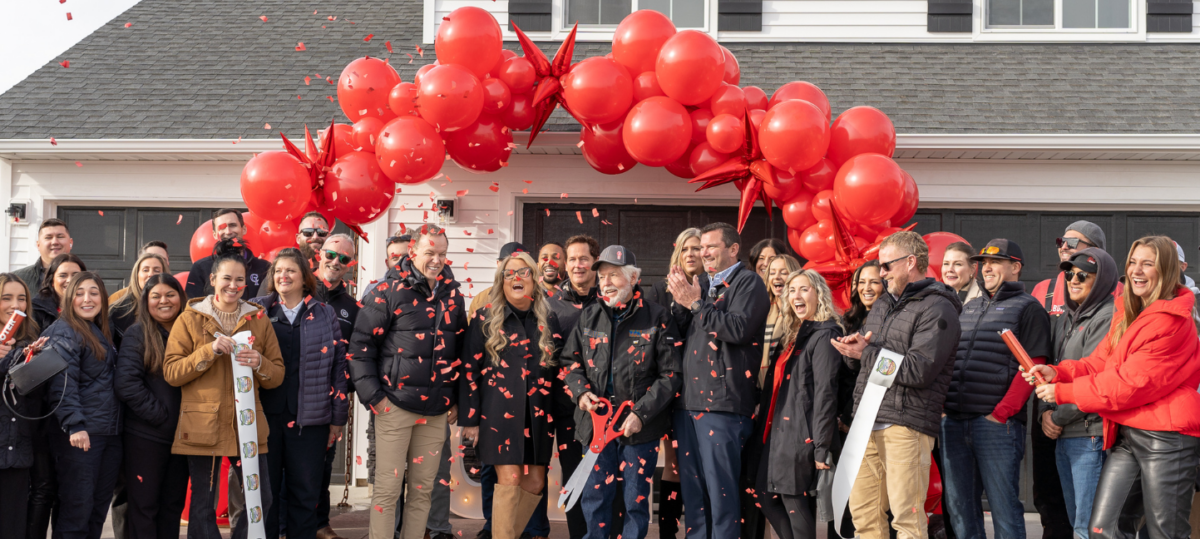 "A lively ribbon-cutting celebration for a new CBH Homes community, featuring a large group of smiling people gathered in front of a newly built white home. The attendees, dressed in warm clothing, cheer as red confetti fills the air. A bold red balloon arch decorates the entrance, while a person in the center holds oversized scissors, symbolizing the official opening of the community. The atmosphere is festive and full of excitement as CBH Homes welcomes new homeowners and neighbors.