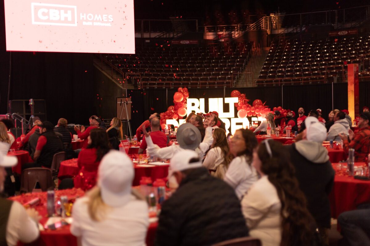 Attendees at the 2025 CBH Homes Company Kickoff event watch an engaging presentation in a large indoor arena, surrounded by red-themed decorations and company branding. A massive screen displays the CBH Homes logo and the phrase ‘Built Different’ is illuminated in bold letters, enhancing the energetic atmosphere. Red confetti fills the air as employees, dressed in company apparel, celebrate and engage with the event. This powerful corporate gathering reflects CBH Homes’ commitment to innovation, team culture, and leadership in the homebuilding industry.