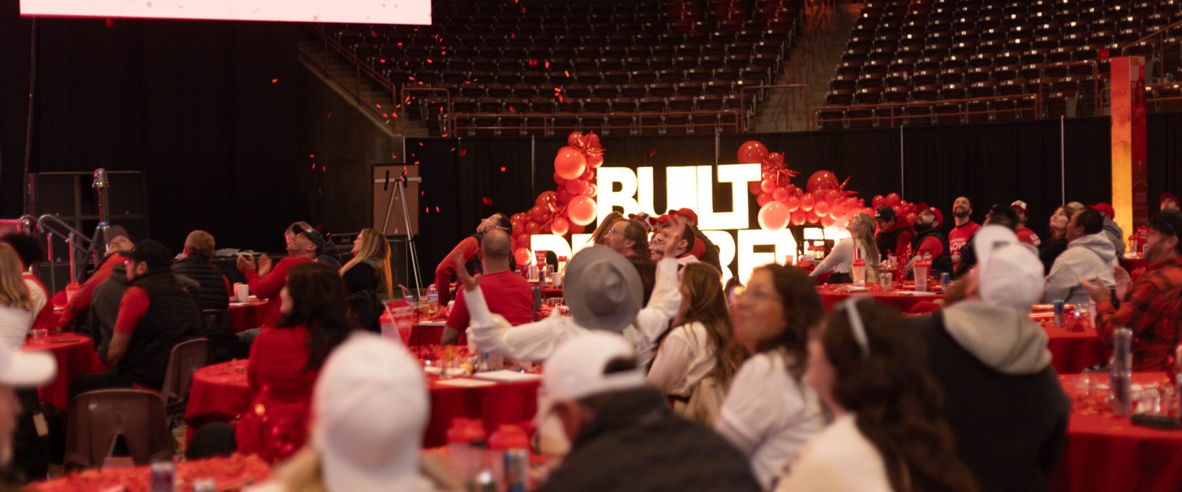 Attendees at the 2025 CBH Homes Company Kickoff event watch an engaging presentation in a large indoor arena, surrounded by red-themed decorations and company branding. A massive screen displays the CBH Homes logo and the phrase ‘Built Different’ is illuminated in bold letters, enhancing the energetic atmosphere. Red confetti fills the air as employees, dressed in company apparel, celebrate and engage with the event. This powerful corporate gathering reflects CBH Homes’ commitment to innovation, team culture, and leadership in the homebuilding industry.