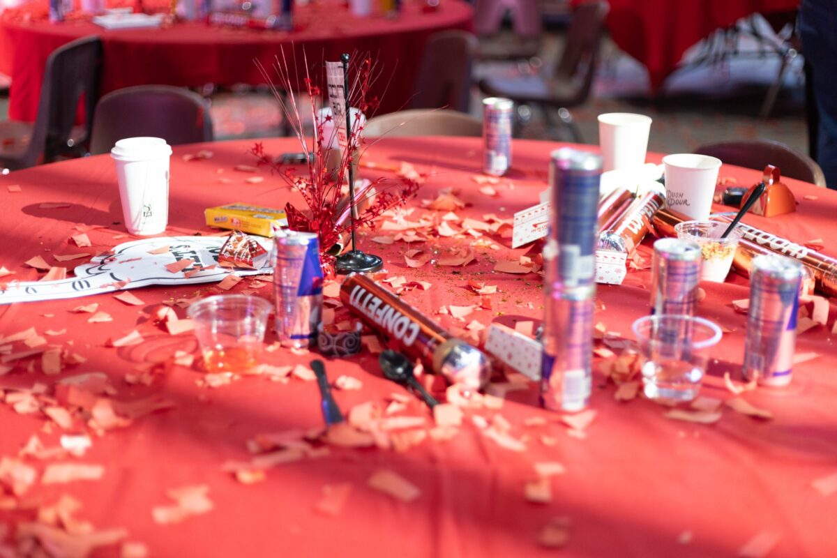 A close-up of a table at the 2025 CBH Homes Company Kickoff event, covered in red decorations, confetti, and party poppers. Empty energy drink cans, disposable coffee cups, snack wrappers, and celebration remnants create a lively post-event atmosphere. The scene captures the excitement and high energy of the CBH Homes corporate gathering, emphasizing a culture of enthusiasm, teamwork, and motivation within the homebuilding industry.