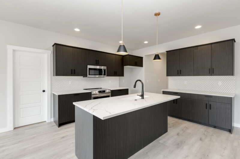 "Modern kitchen in the Vallejo 2700 floor plan by CBH Homes – featuring dark espresso cabinets, a stunning white quartz island with waterfall edge, hex tile backsplash, and sleek black fixtures in the Prominence community.