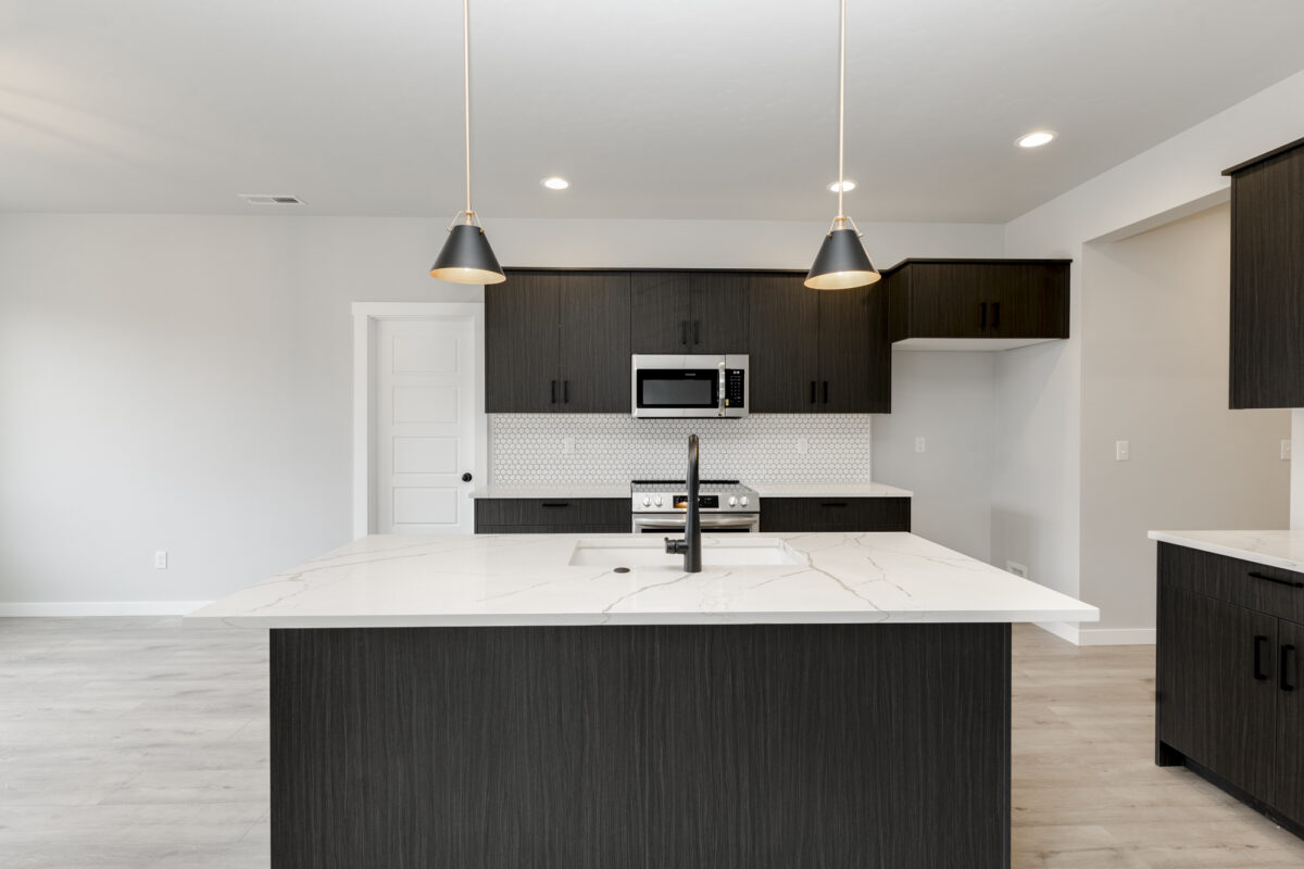 Straight-on view of the Vallejo 2700 kitchen by CBH Homes – showcasing a bold black island with white quartz countertops, modern black pendant lighting, dark wood cabinetry, and a stylish white hex backsplash in the Prominence community.