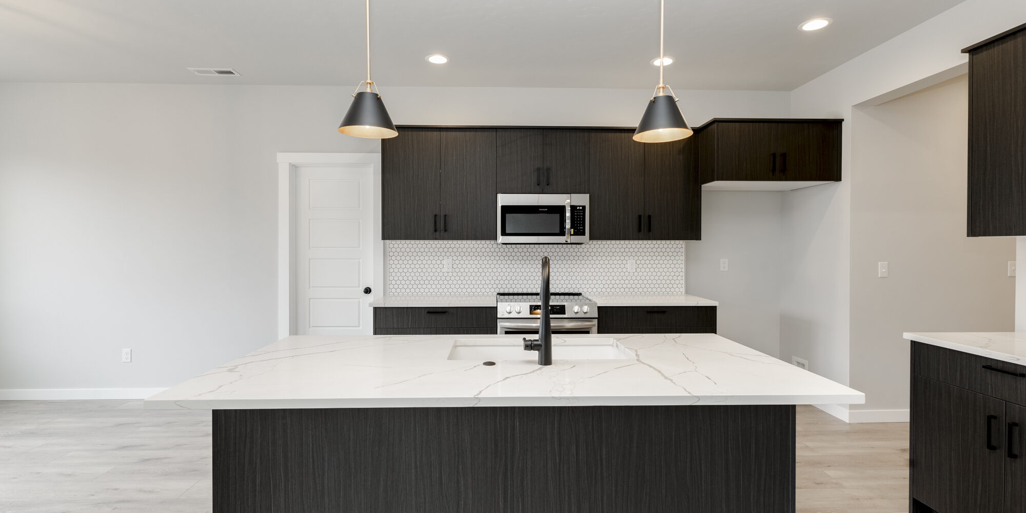 Straight-on view of the Vallejo 2700 kitchen by CBH Homes – showcasing a bold black island with white quartz countertops, modern black pendant lighting, dark wood cabinetry, and a stylish white hex backsplash in the Prominence community.