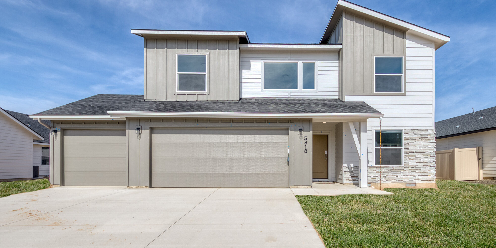 Modern two-story home with a three-car garage, light gray and white exterior, and stone accents. The house features a covered front porch, large windows, and a well-maintained lawn under a bright blue sky.